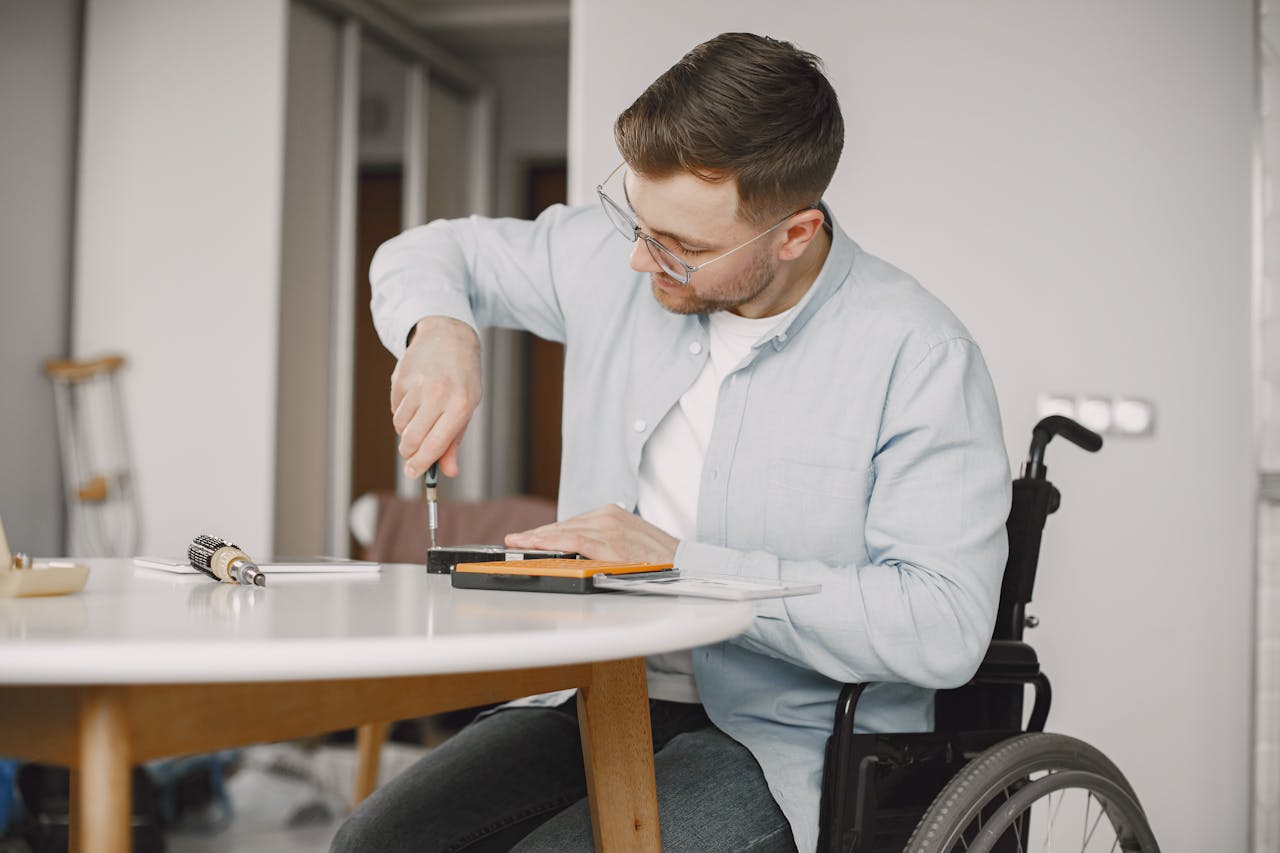 A man in a wheelchair uses a screwdriver at a home desk, engaging in repair work.