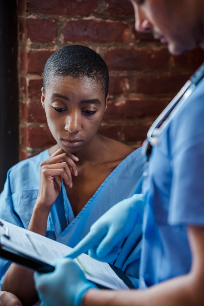 Thoughtful African American woman patient with short hair in medical robe looking at clipboard with diagnosis while talking with doctor in hallway of hospital